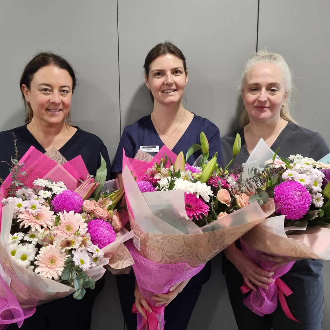 Three female nurses wearing blue scrubs smile at the camera while holding large bouquets of multicoloured flowers.